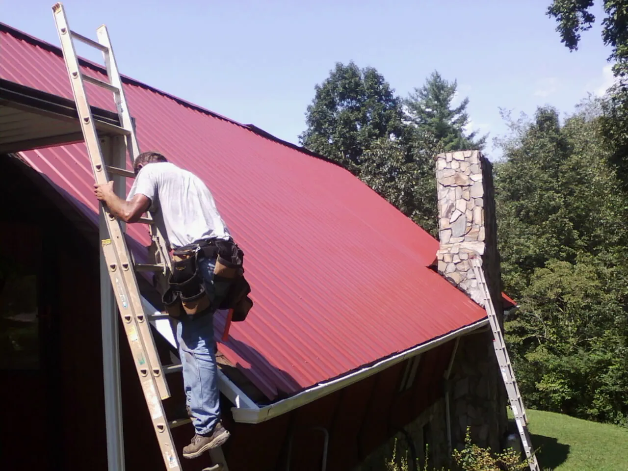 Roofer on a ladder beside a red metal roof and stone chimney — Soffit & Fascia Repair by Roofing Brothers in Succasunna